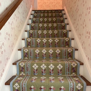 Puede incluir: Una escalera con corredores de alfombra con estampado floral verde oliva. La alfombra presenta un patrón repetitivo de flores blancas, moradas y rosas. Las escaleras tienen escalones de madera y contrahuellas blancas, con varillas de latón. Las paredes están cubiertas de papel pintado floral.