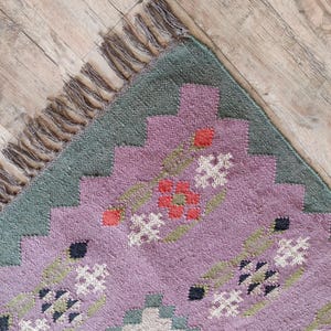 Pode incluir: Um close-up de um tapete estampado em roxo e verde com uma borda de franjas. O tapete tem um design floral com detalhes em vermelho, branco e preto.