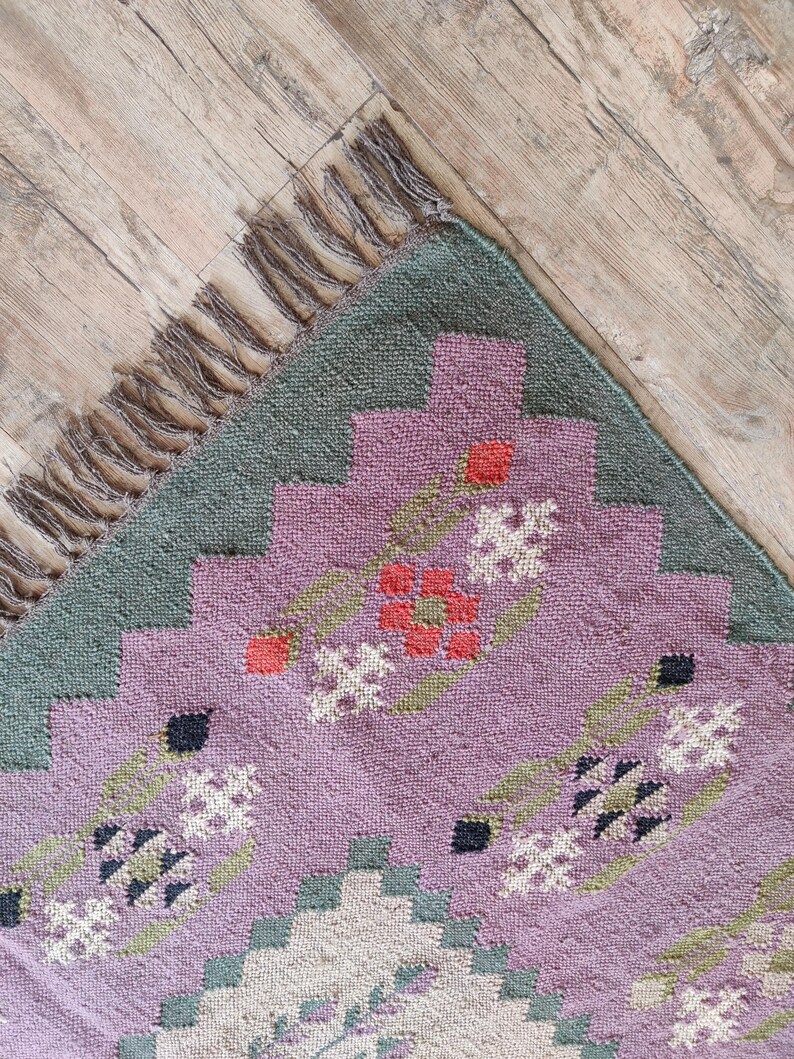 Pode incluir: Um close-up de um tapete estampado em roxo e verde com uma borda de franjas. O tapete tem um design floral com detalhes em vermelho, branco e preto.