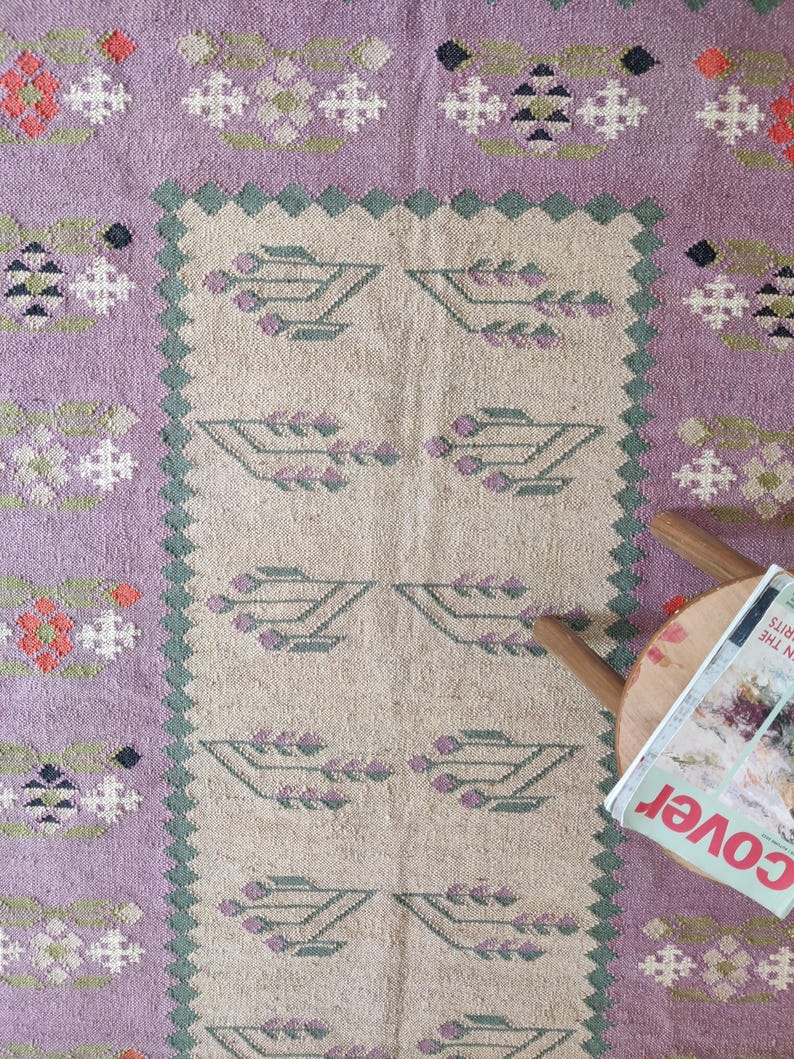 Pode incluir: Um tapete tecido &agrave; m&atilde;o com um fundo roxo claro e um centro bege. O centro apresenta um padr&atilde;o repetitivo de motivos florais estilizados em tons de verde e roxo. O tapete tem uma borda com franjas.