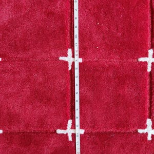May include: A close-up of a red rug with a white plus sign pattern. The rug is approximately 11 inches long. The rug has a soft, textured surface. The plus signs are evenly spaced across the rug.