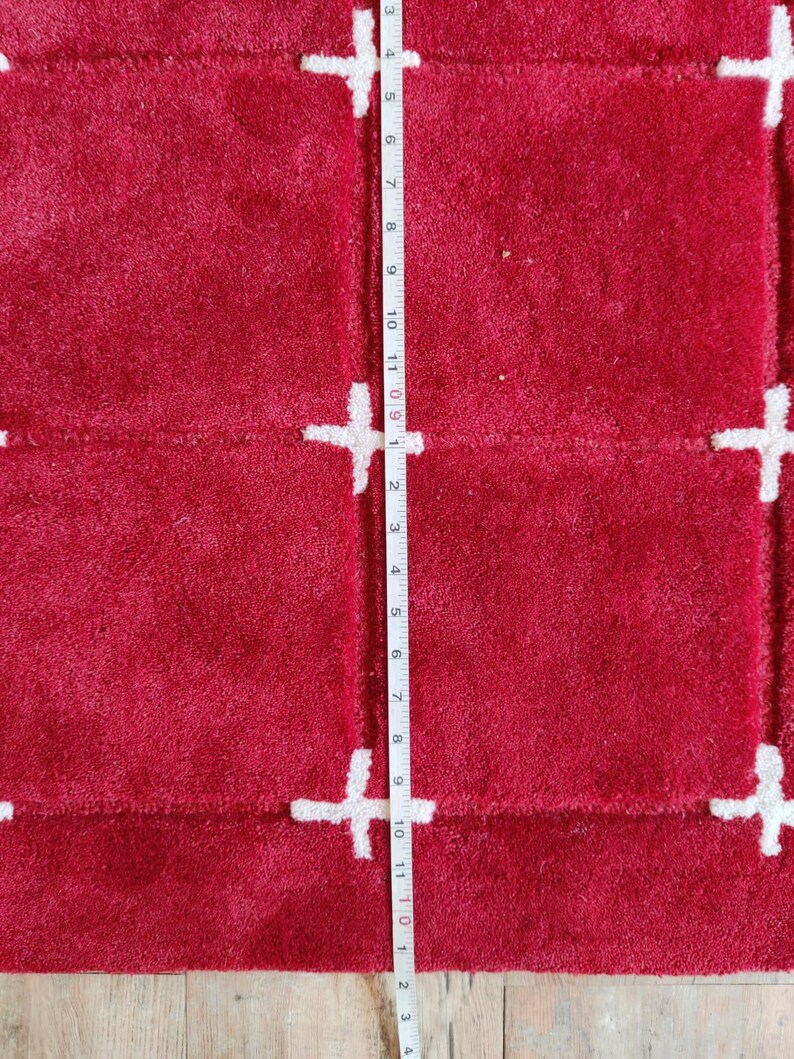 May include: A close-up of a red rug with a white plus sign pattern. The rug is approximately 11 inches long. The rug has a soft, textured surface. The plus signs are evenly spaced across the rug.