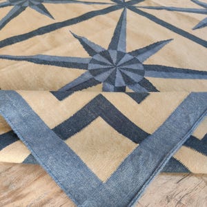 May include: A beige and blue geometric patterned rug. The rug features a repeating starburst design in shades of blue against a beige background. The border of the rug is a solid blue with a zig-zag pattern.