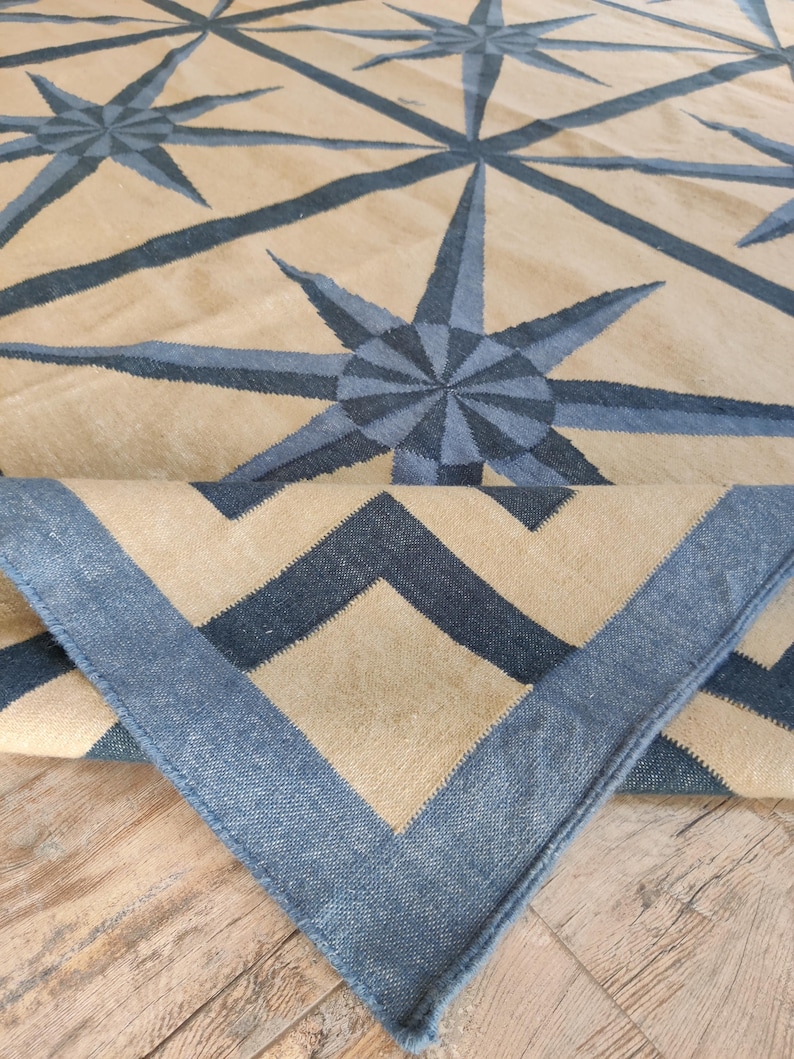 May include: A beige and blue geometric patterned rug. The rug features a repeating starburst design in shades of blue against a beige background. The border of the rug is a solid blue with a zig-zag pattern.