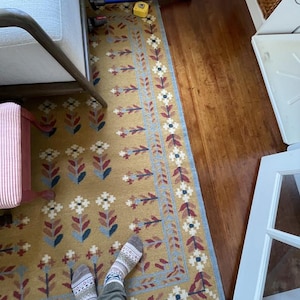 May include: Mustard yellow rectangular rug with a floral pattern in white, blue, and burgundy. The rug has a blue border with white flowers and a fringe along the bottom edge. The rug is placed on a hardwood floor.