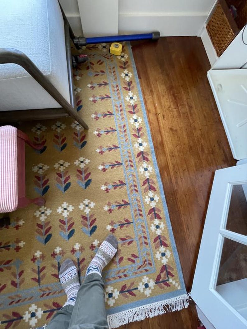 May include: Mustard yellow rectangular rug with a floral pattern in white, blue, and burgundy. The rug has a blue border with white flowers and a fringe along the bottom edge. The rug is placed on a hardwood floor.