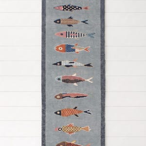 May include: A rectangular runner rug in a muted blue-gray tone featuring a vertical arrangement of eleven stylized fish in various colors and geometric patterns. The rug is framed by a dark blue border and finished with short fringe at both ends.