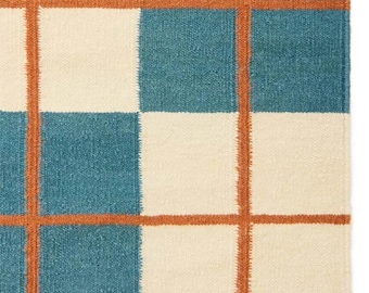 May include: Close-up of a woven rug with a grid pattern. The rug features alternating squares of cream and teal blue, with rust-colored lines forming the grid. The texture of the rug is visible, suggesting a natural fiber material.