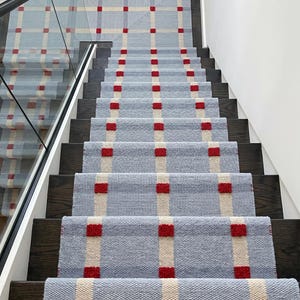 May include: A stair runner featuring a light blue woven base with a grid pattern of cream-colored vertical and horizontal stripes. Small, raised, bright red square accents are positioned at each intersection of the cream-colored stripes.