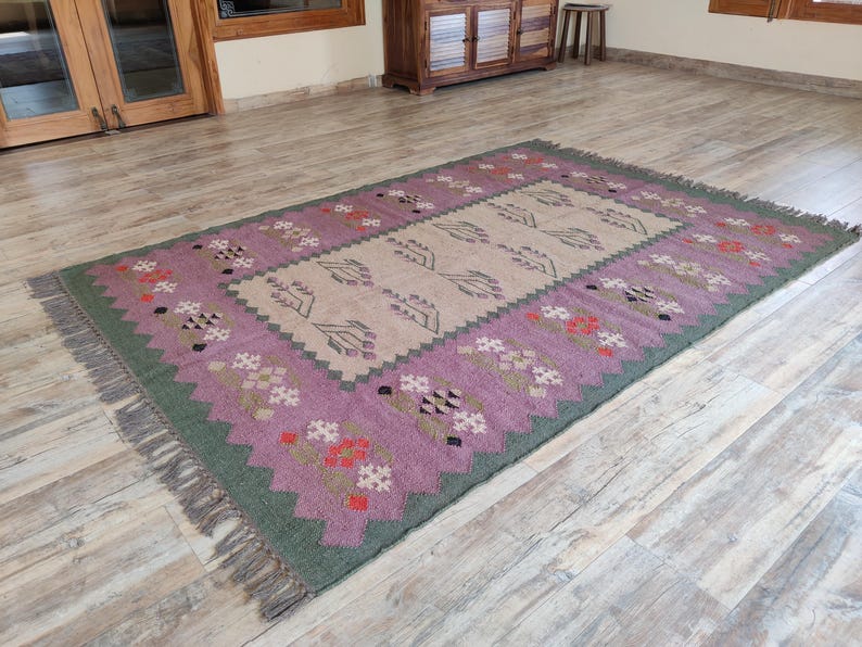 Pode incluir: Um tapete tecido &agrave; m&atilde;o com um padr&atilde;o geom&eacute;trico rosa e verde. O tapete tem um centro bege com um desenho floral. O tapete tem franjas em todos os lados.