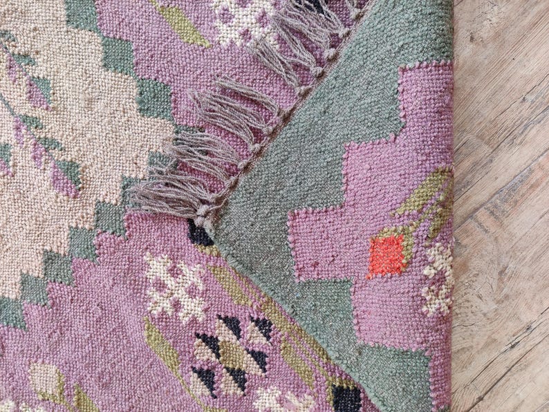Pode incluir: Um close-up de um tapete tecido rosa, verde e branco com uma borda de franja. O tapete tem um padr&atilde;o geom&eacute;trico e &eacute; feito de um material macio e texturizado.
