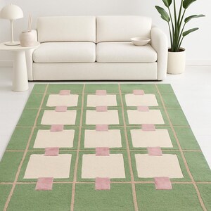 May include: A rectangular green area rug with a geometric design. The rug features a grid pattern with cream-colored rectangles and pink squares. The rug is placed in front of a white sofa.
