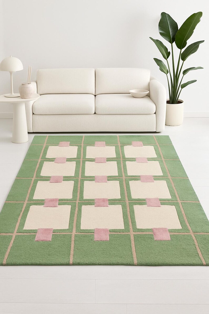 May include: A rectangular green area rug with a geometric design. The rug features a grid pattern with cream-colored rectangles and pink squares. The rug is placed in front of a white sofa.
