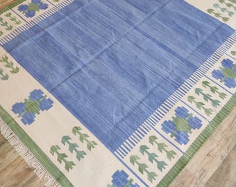 May include: A rectangular area rug featuring a large solid blue center panel surrounded by a cream-colored border. The border displays repeating blue and green floral motifs, striped accents, and green leaf patterns, finished with white fringe.
