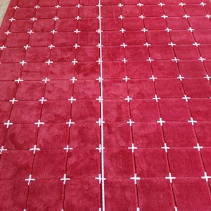 May include: A large, rectangular red rug with a repeating pattern of white plus signs. The rug appears to be made of a soft, plush material. A measuring tape is laid across the center of the rug.