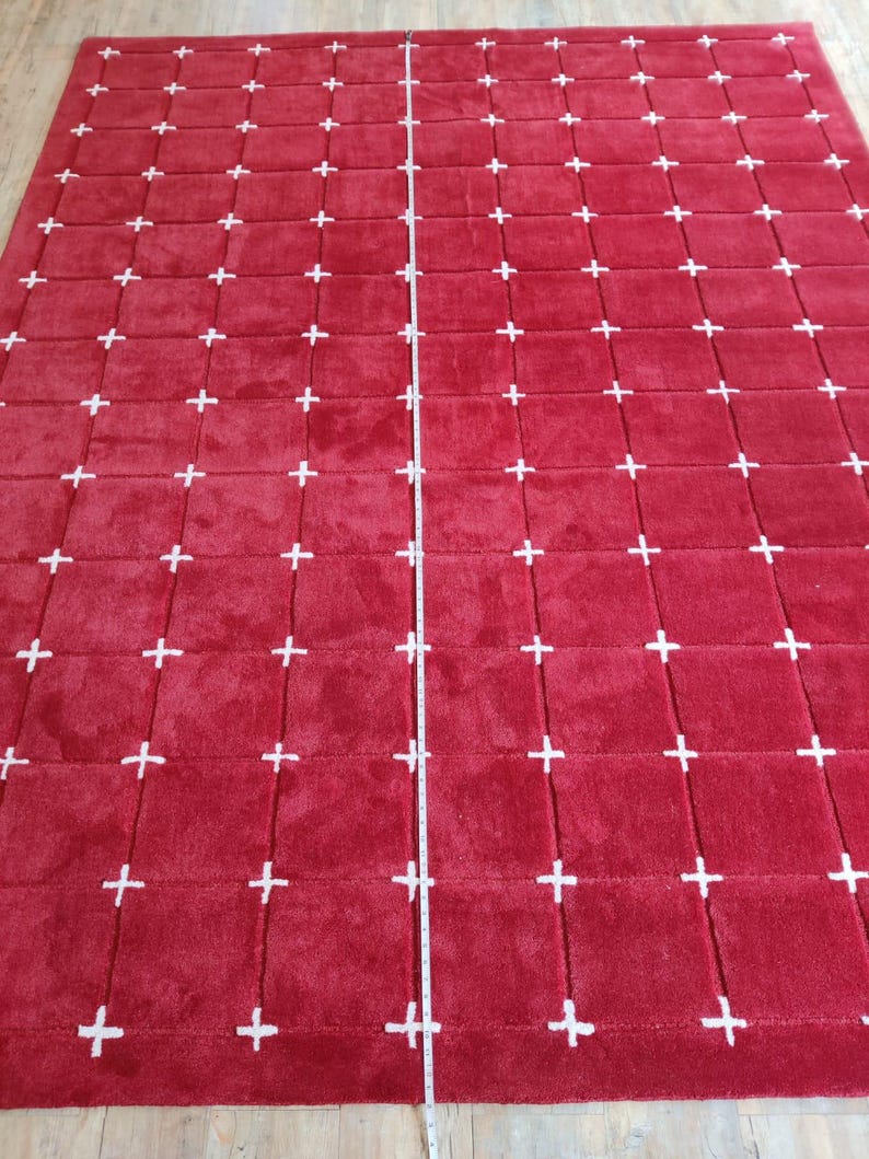 May include: A large, rectangular red rug with a repeating pattern of white plus signs. The rug appears to be made of a soft, plush material. A measuring tape is laid across the center of the rug.