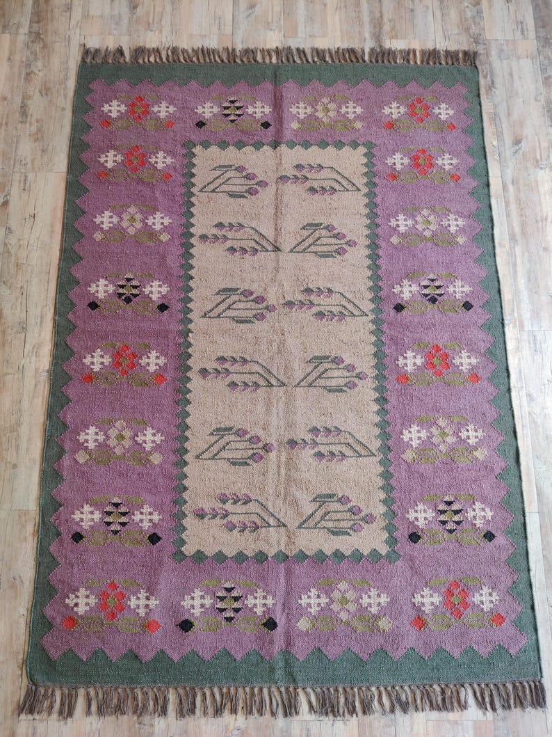 Pode incluir: Um tapete tecido &agrave; m&atilde;o com um padr&atilde;o geom&eacute;trico em tons de verde, roxo e bege. O tapete tem uma borda de franjas.
