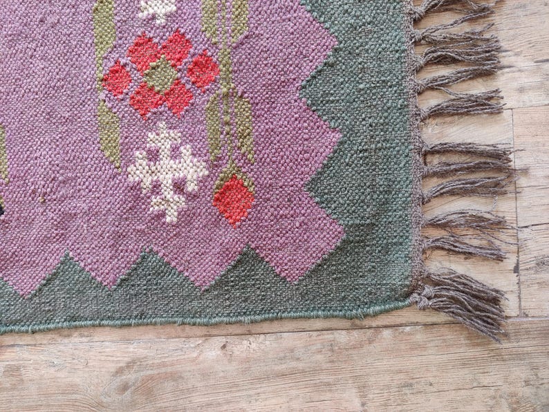 Pode incluir: Um tapete tecido de cor roxa e verde com um padr&atilde;o floral e franjas. O tapete tem um design geom&eacute;trico com uma flor rosa no centro.