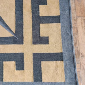 May include: A rectangular rug with a geometric design. The rug features a tan background with a dark blue border and dark blue geometric shapes. The rug is made of a woven material and is placed on a wooden floor.