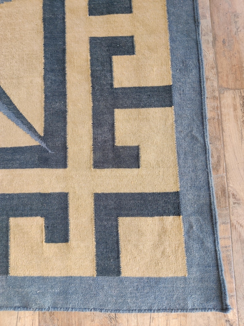 May include: A rectangular rug with a geometric design. The rug features a tan background with a dark blue border and dark blue geometric shapes. The rug is made of a woven material and is placed on a wooden floor.