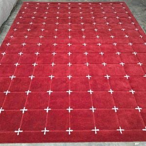 May include: A large, rectangular red rug with a grid pattern of white plus signs. The rug has a plush texture and is likely made of a soft material. The design is simple yet eye-catching, suitable for various interior styles.