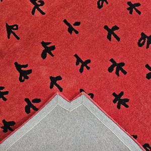 May include: A red rug with a repeating pattern of black abstract shapes. The rug's underside is visible, showing a gray backing with a stitched border. The rug's texture appears soft and plush, suitable for a living space.