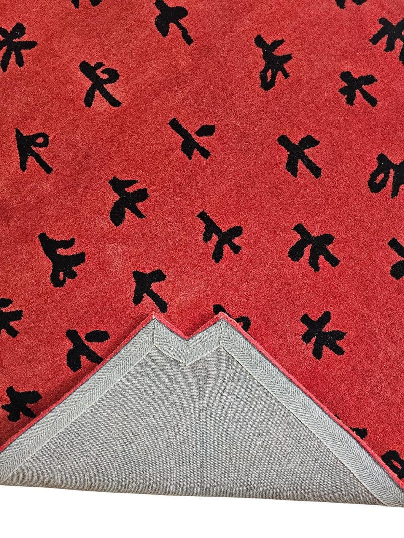 May include: A red rug with a repeating pattern of black abstract shapes. The rug's underside is visible, showing a gray backing with a stitched border. The rug's texture appears soft and plush, suitable for a living space.