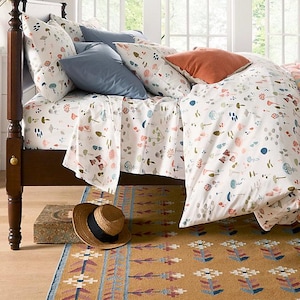 May include: A bed with a white duvet cover and matching sheets, patterned with colorful mushrooms and other botanical elements. The bed is made with blue and orange pillows. A straw hat and a patterned rug are on the floor.