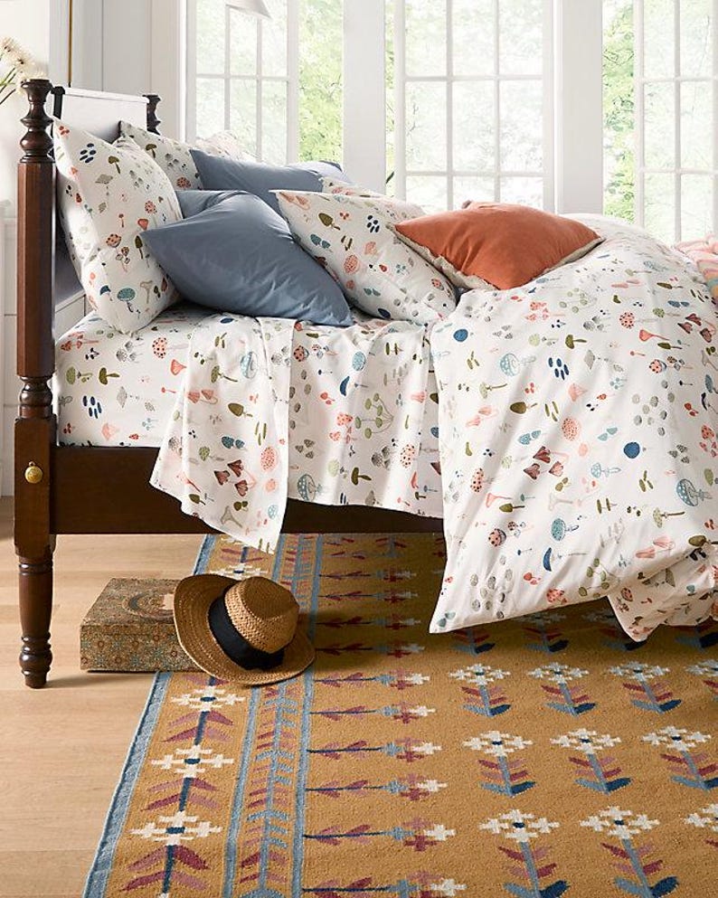 May include: A bed with a white duvet cover and matching sheets, patterned with colorful mushrooms and other botanical elements. The bed is made with blue and orange pillows. A straw hat and a patterned rug are on the floor.