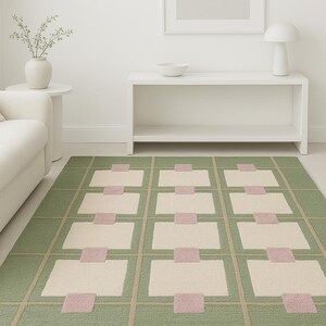 May include: A rectangular area rug with a geometric pattern. The rug features a green background with a grid of beige squares and pink rectangles. The rug is placed on a light-colored floor, and a white sofa is visible in the background.