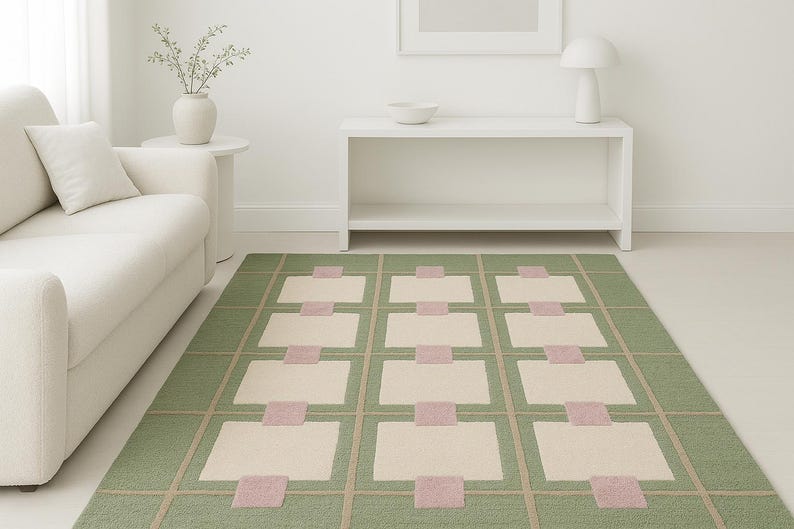 May include: A rectangular area rug with a geometric pattern. The rug features a green background with a grid of beige squares and pink rectangles. The rug is placed on a light-colored floor, and a white sofa is visible in the background.