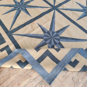 May include: A beige and blue patterned rug with a geometric design. The rug features a repeating starburst pattern in shades of blue, with a border of geometric shapes. The rug is partially rolled up, revealing the underside.