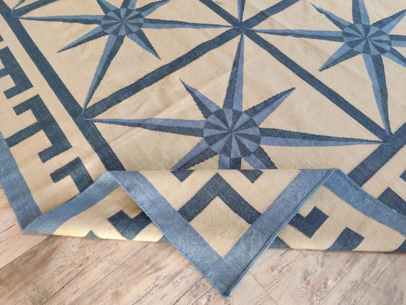 May include: A beige and blue patterned rug with a geometric design. The rug features a repeating starburst pattern in shades of blue, with a border of geometric shapes. The rug is partially rolled up, revealing the underside.