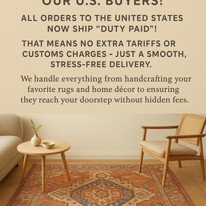 May include: A rectangular area rug with a central blue diamond pattern on an orange background, featuring intricate borders. The rug is placed in a room with a light wood chair, a small round table with a vase and bowl, and a portion of a sofa.