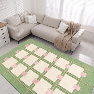 May include: A rectangular area rug with a green background and a grid pattern. The grid features beige lines and squares, with pink accents. The rug is placed in a living room setting with a beige sectional sofa and a white dresser.