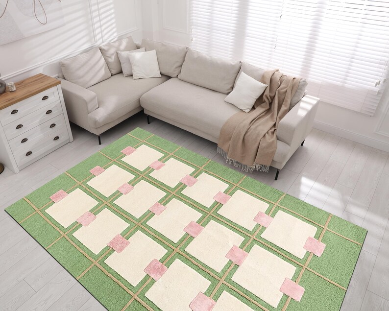 May include: A rectangular area rug with a green background and a grid pattern. The grid features beige lines and squares, with pink accents. The rug is placed in a living room setting with a beige sectional sofa and a white dresser.