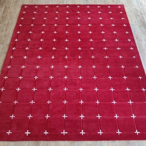 May include: A large, rectangular red rug with a grid pattern of white plus signs. The rug is made of a soft material and is placed on a wooden floor.