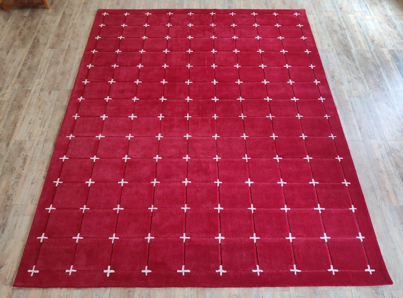 May include: A large, rectangular red rug with a grid pattern of white plus signs. The rug is made of a soft material and is placed on a wooden floor.