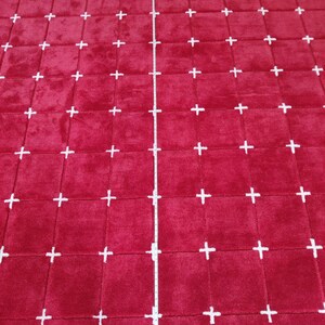 May include: A large, rectangular red rug with a grid pattern of white plus signs. The rug appears to be made of a soft, plush material. A measuring tape is placed vertically on the rug.