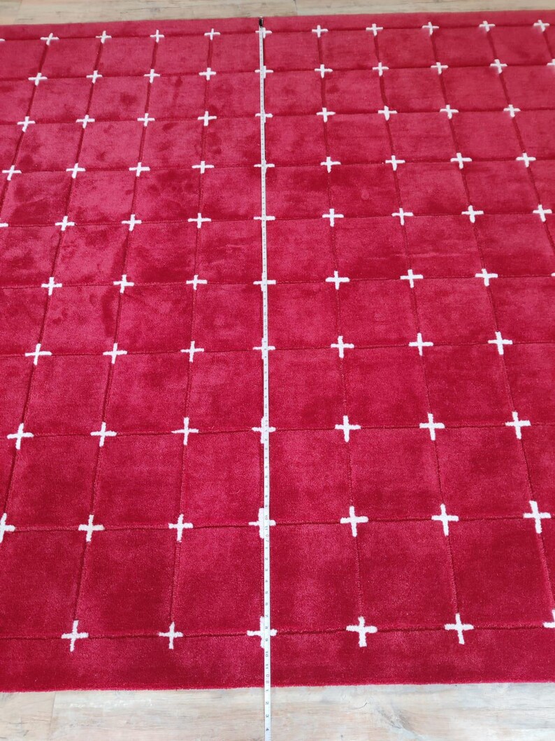 May include: A large, rectangular red rug with a grid pattern of white plus signs. The rug appears to be made of a soft, plush material. A measuring tape is placed vertically on the rug.