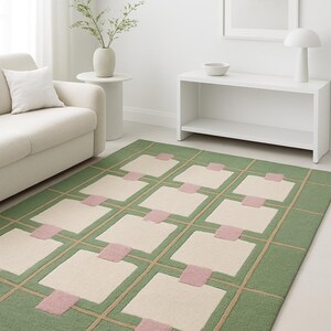 May include: A rectangular area rug with a geometric pattern. The rug features a green border and background, with beige squares and pink accents. Beige lines create a grid pattern over the green background.