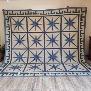 May include: Large rectangular rug with a cream background and a repeating pattern of blue eight-pointed stars. The rug has a blue border with a geometric design. The rug is displayed on a wooden floor.