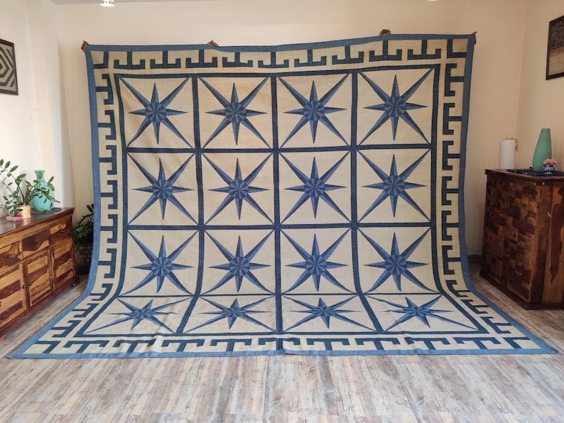 May include: Large rectangular rug with a cream background and a repeating pattern of blue eight-pointed stars. The rug has a blue border with a geometric design. The rug is displayed on a wooden floor.