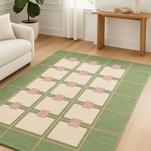 May include: A rectangular area rug with a geometric design. The rug features a green border and a grid pattern of cream-colored squares, accented with pink rectangles. The rug is placed on a hardwood floor.