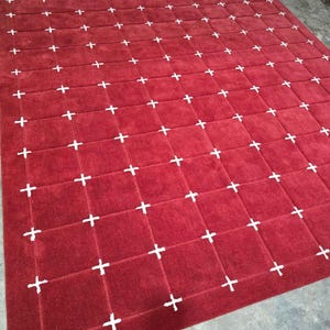 May include: A large, rectangular red rug with a grid pattern of white plus signs. The rug has a plush texture and is suitable for a living room or bedroom. The rug is made of a soft material.