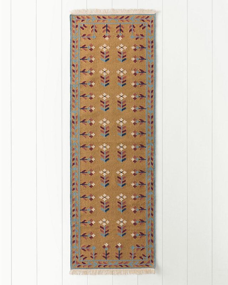 May include: A rectangular, mustard-colored runner rug with a repeating floral pattern in shades of red, blue, and white. The rug has a blue border and fringed edges. The design is symmetrical and traditional.