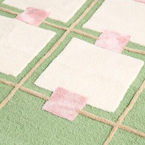 May include: A close-up of a rug with a geometric pattern. The rug features a light green background with a grid of beige lines. Cream-colored squares are interspersed with small pink squares. The rug has a soft, textured appearance.