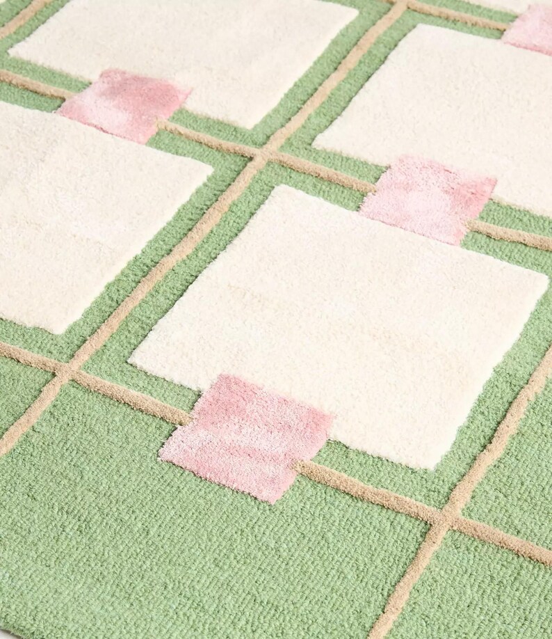 May include: A close-up of a rug with a geometric pattern. The rug features a light green background with a grid of beige lines. Cream-colored squares are interspersed with small pink squares. The rug has a soft, textured appearance.