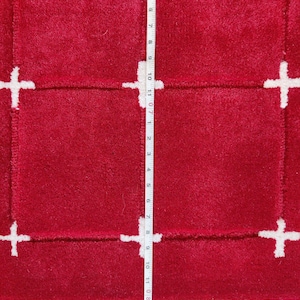 May include: A close-up of a red rug with a white cross pattern. The rug has a plush texture and a rectangular design. A measuring tape is placed vertically along the rug, showing measurements in inches.