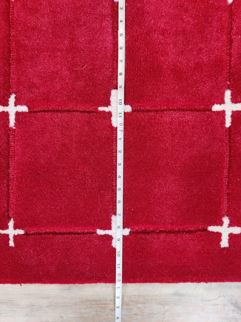 May include: A close-up of a red rug with a white cross pattern. The rug has a plush texture and a rectangular design. A measuring tape is placed vertically along the rug, showing measurements in inches.